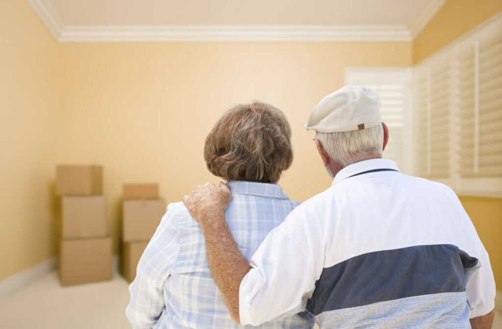 An elderly couple sitting side-by-side looking at their packed boxes in an empty room as they move into a senior living community.