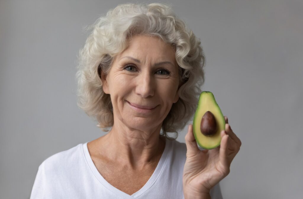 An older adult woman holding half an avocado with her left hand while smiling