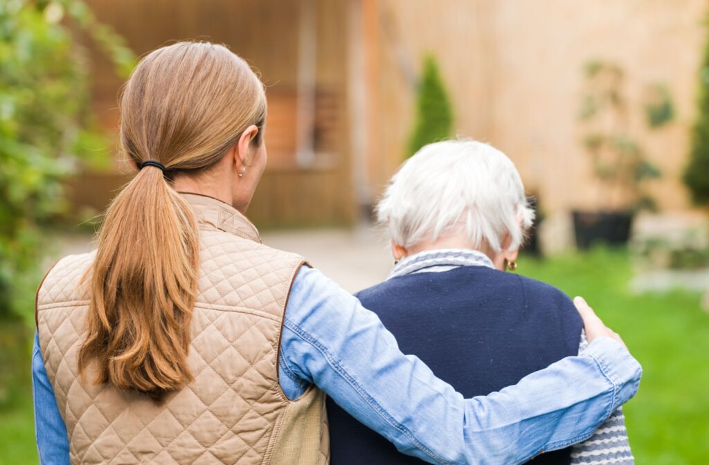 A caregiver and a senior with dementia walk side by side outdoors, with the caregiver gently supporting the senior.