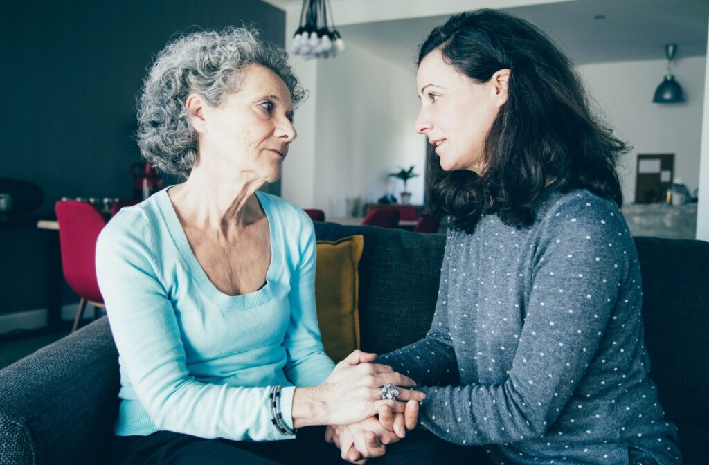 An adult holds their senior parent’s hands as they have a serious conversation in their living room about assisted living