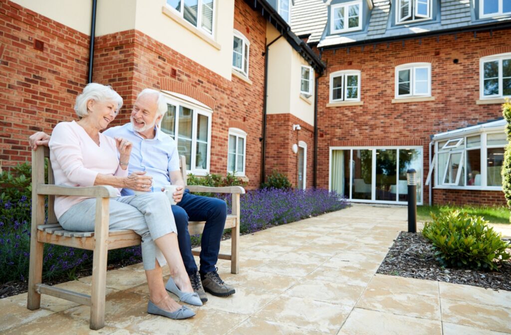 An older couple sitting on a bench with cups of coffee in the courtyard of their new senior living community.