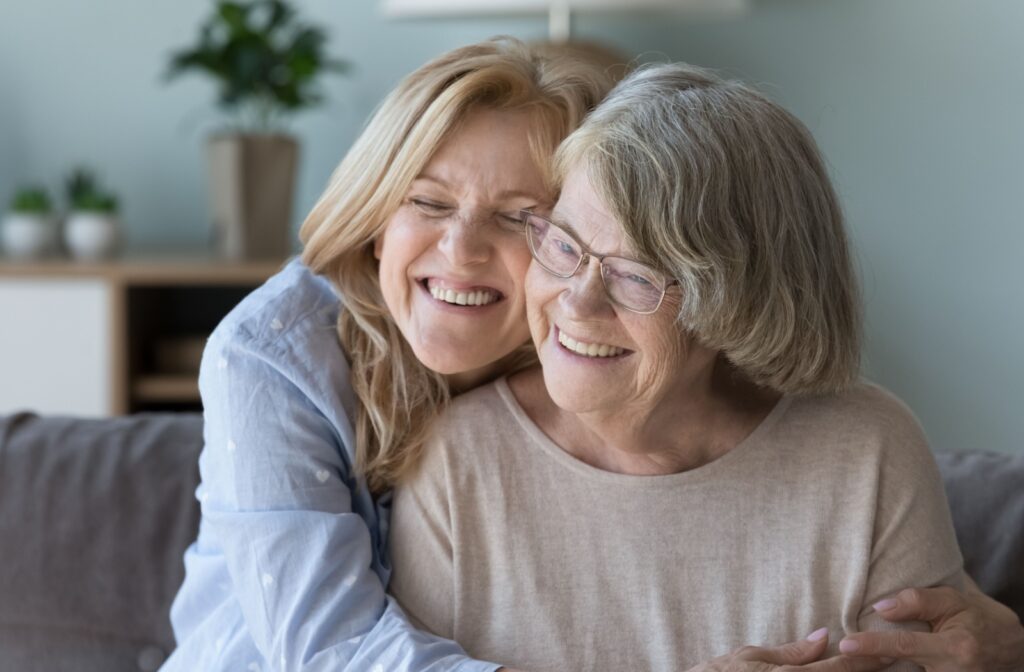 An older adult smiles while their adult child laughs and hugs them while they sit together on a couch