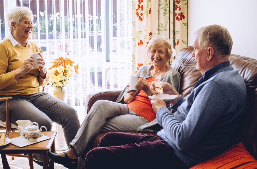 3 older adults enjoy pastries and coffee on a leather couch while sitting beside a warm sunlit window in the lounge of their assisted living community