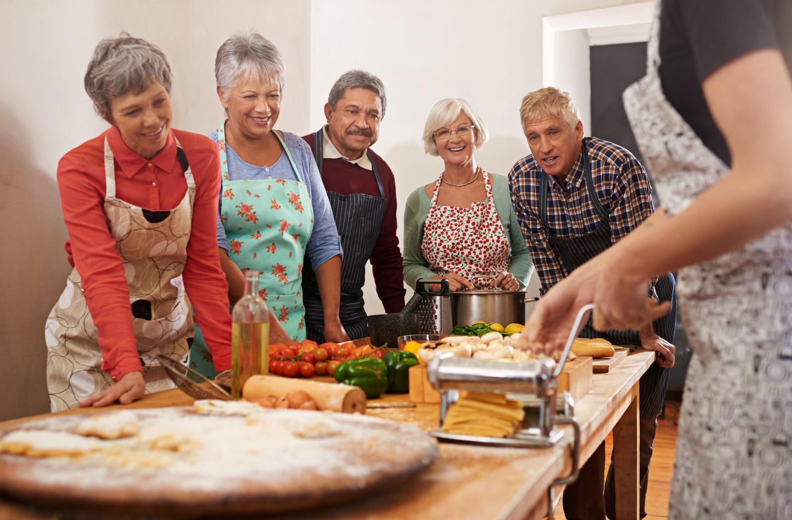 A group of seniors learning how to make pasta from a professional chef in the kitchen.